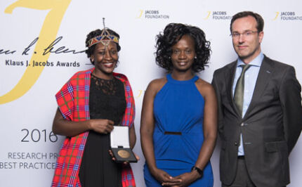 Two women and a man stand in front of a Jacobs Foundation banner holding their best practice prize trophy