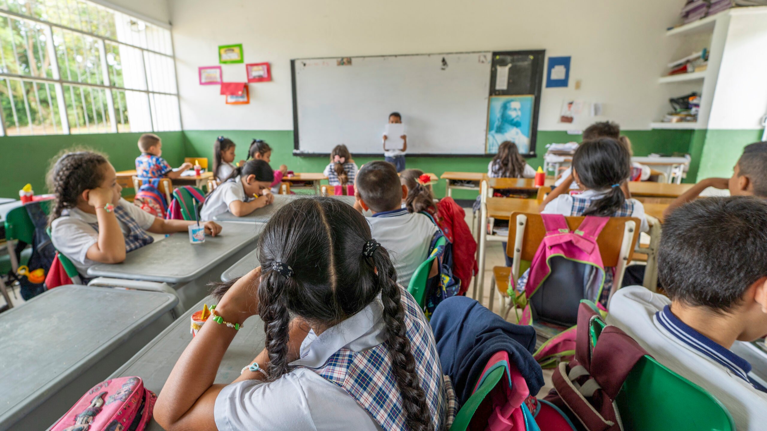 Primary school students sitting at desks in a classroom, listening as a child presents in front of a whiteboard.